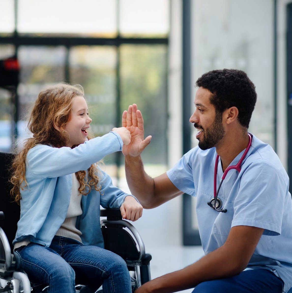 Arzt gibt fröhlichem Mädchen im Rollstuhl ein High Five im Krankenhausflur