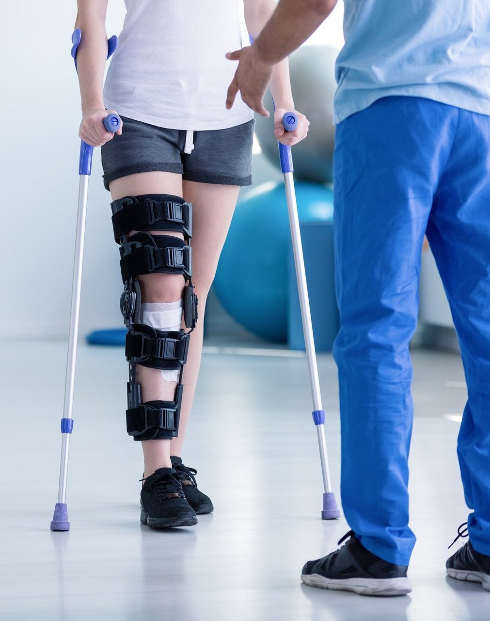 Eine Person mit Beinorthese und Gehstützen wird in einer Rehaklinik von einem Therapeuten beim Gehen unterstützt.