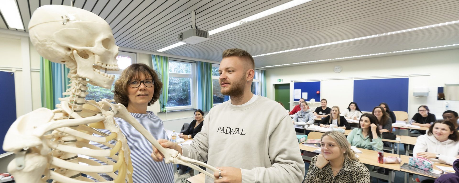 Das Bild zeigt ein Klassenzimmer mit Schülern. Ein Schüler steht vorne und bewegt den Arm eines Skeletts. Neben ihm steht eine Lehrerin.