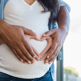 Ein Paar formt zusammen mit ihren Händen ein Herz und halten es vor den Bauch der schwangeren Frau.