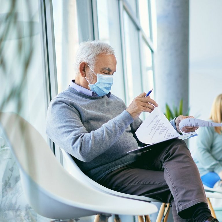 In einem Wartebereich eines Krankenhauses oder einer Arztpraxis sitzen mehrere Personen mit Masken, während ein älterer Mann ein Formular ausfüllt und zwei medizinische Fachkräfte miteinander sprechen.