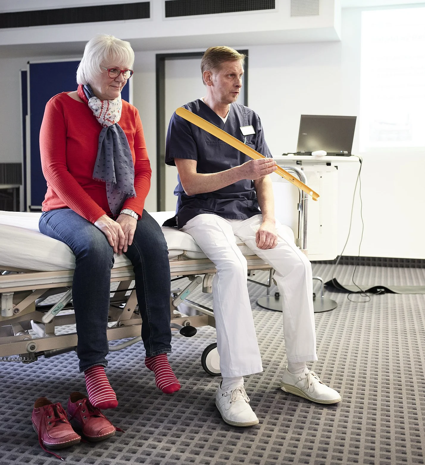 Therapeut demonstriert einer älteren Frau den Umgang mit einem Therapiegurt auf einer Untersuchungsliege.