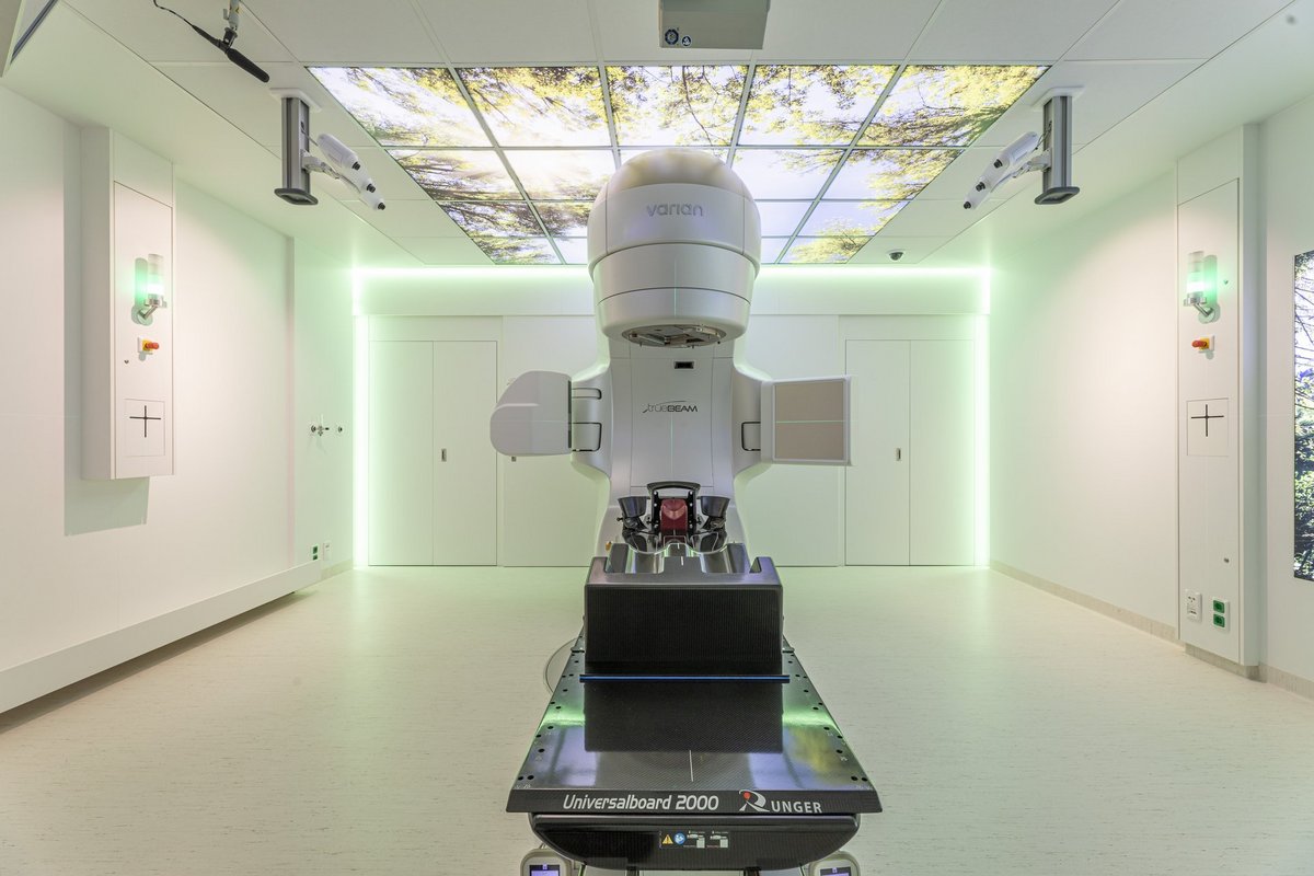 Ein Gerät zur Strahlentherapie steht in einem hellen Raum. An der Decke und den Wänden hängen Landschaftsbilder.