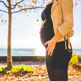 Eine schwangere Frau steht im Herbst auf einem Feld und fasst ihren Bauch an.