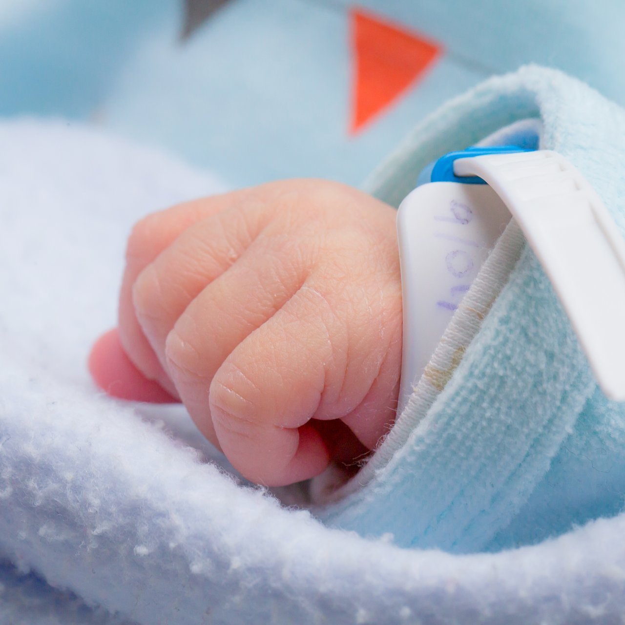 Nahaufnahme einer kleinen Babyhand, die in einem dicken Wolloberteil steckt. An der Hand hängt ein Krankenhausband.