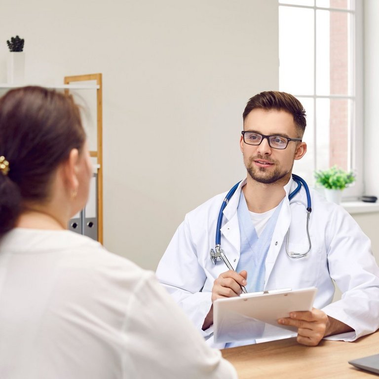 Ein Arzt mit Stethoskop und Klemmbrett spricht mit einer Patientin in einem Büro.