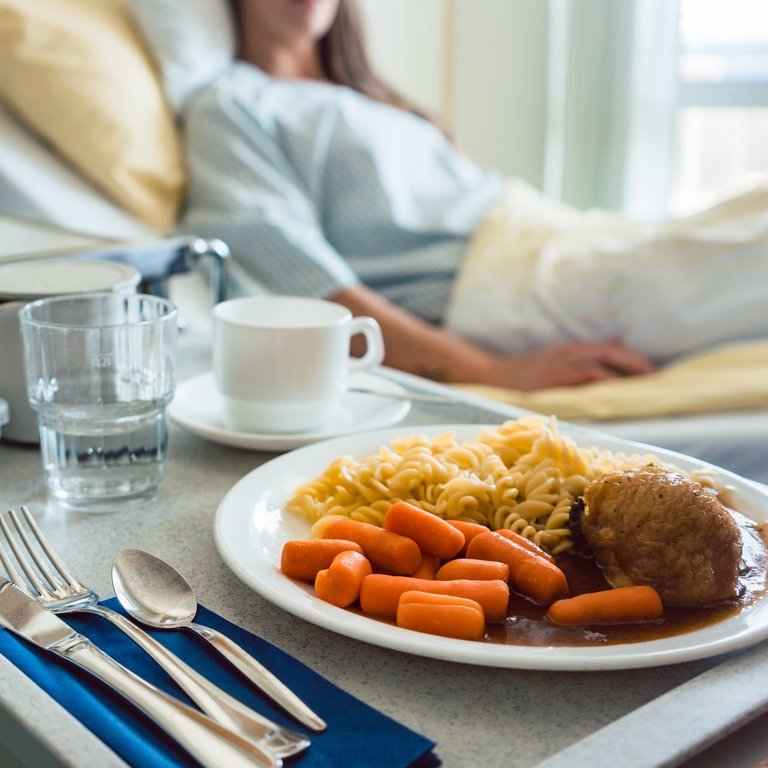 Essen wird einem Patienten im Krankenhaus ans Bett serviert. Der Fokus liegt auf dem Essen, das aus Karotten, Nudeln, Fleisch und dunkler Sauce besteht.