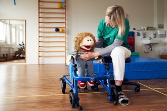 Therapeutin unterstützt eine Handpuppe bei einer Übung mit einem Gehtrainer in einem Therapieraum.