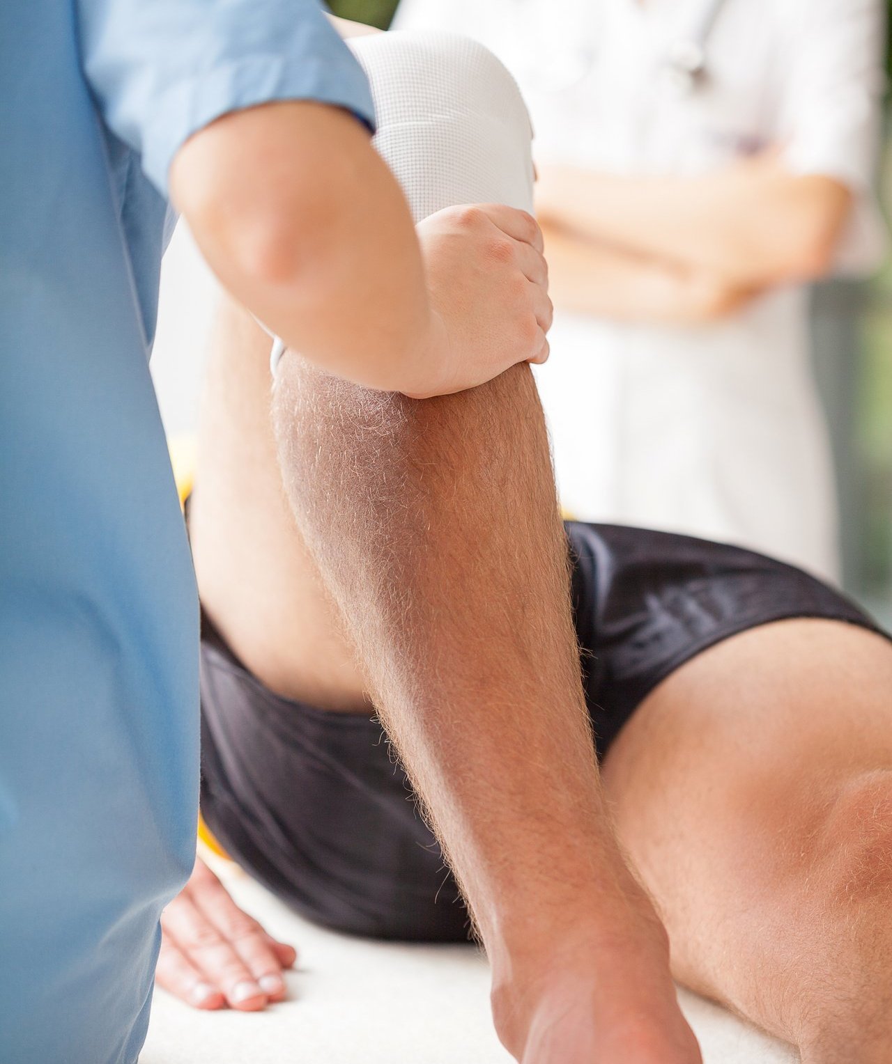 Physiotherapeutin mobilisiert bandagiertes Knie eines männlichen Patienten während Rehabilitationsbehandlung