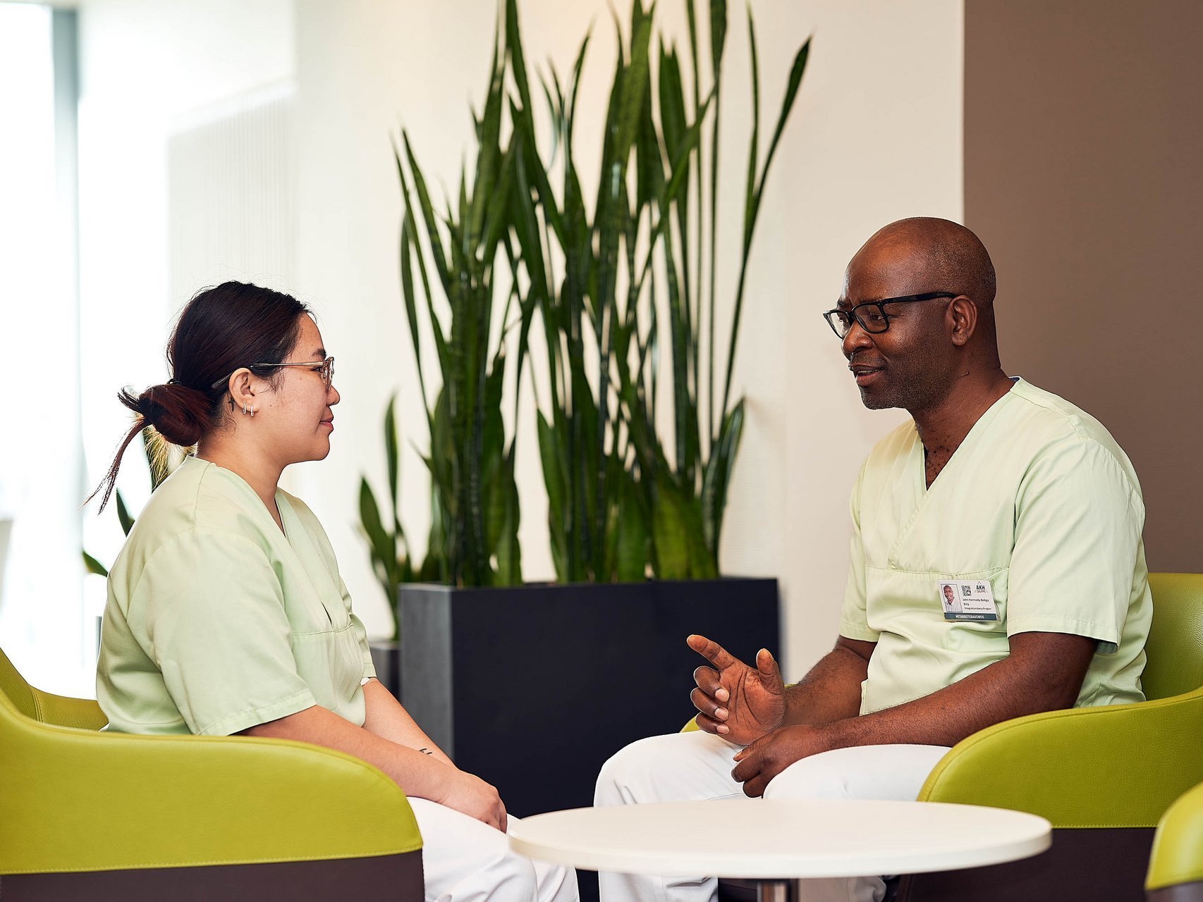Zwei Pflegekräfte in grüner Kleidung sitzen sich in einer modernen, hellen Sitzecke gegenüber und unterhalten sich freundlich. Im Hintergrund steht eine große Zimmerpflanze.