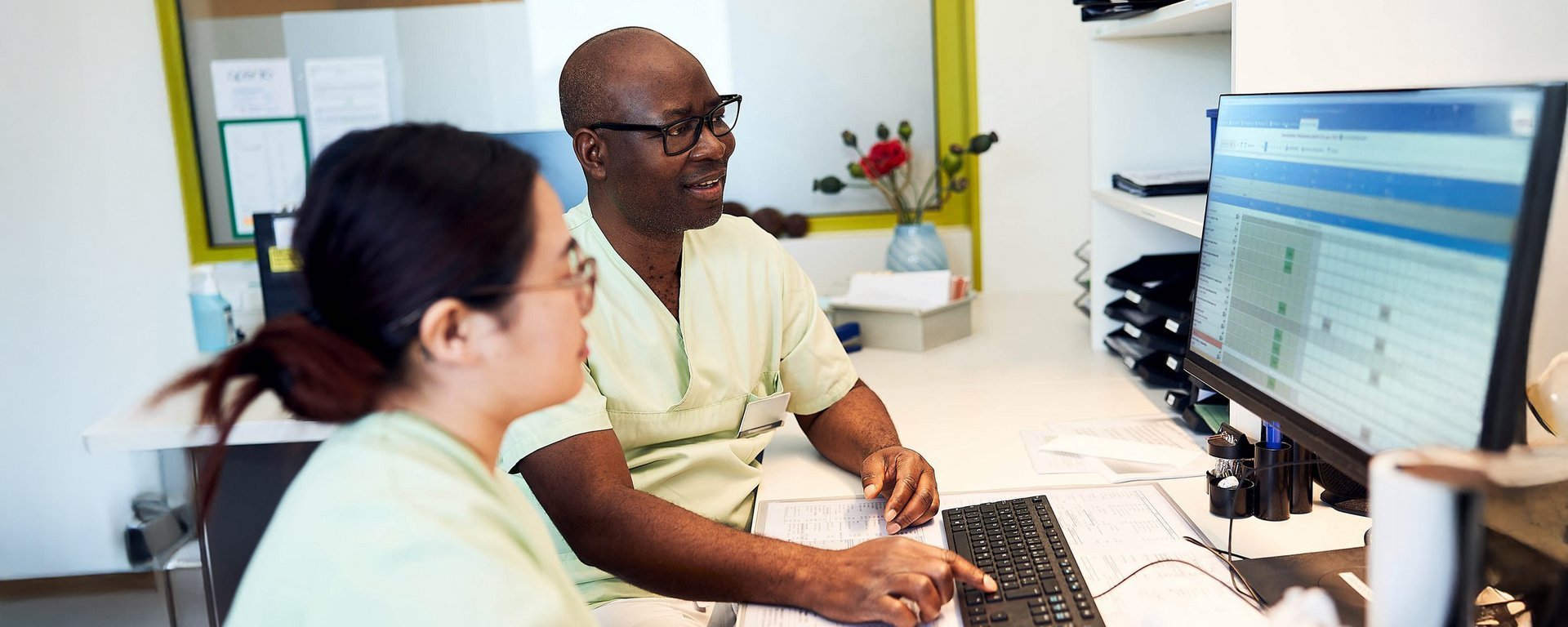 Zwei Pfleger (eine Frau, ein Mann) blicken auf einen Computerbildschirm auf dem ein Programm geöffnet ist. Das Programm zeigt eine Tabelle, in der verschiedene Bereiche farbig markiert sind.