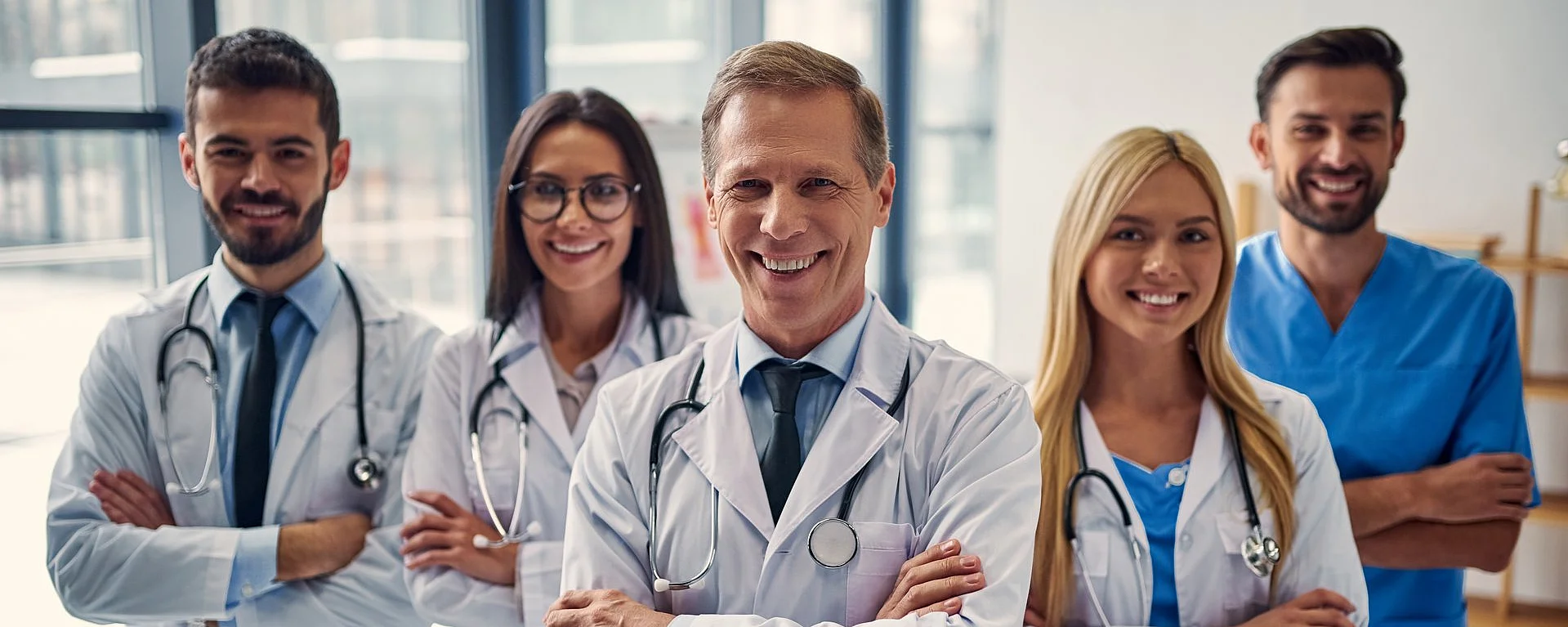 Lächelndes medizinisches Team mit Ärzten und Ärztinnen in weißem Kittel und Pflegekraft in blauer Kleidung im Klinikflur.