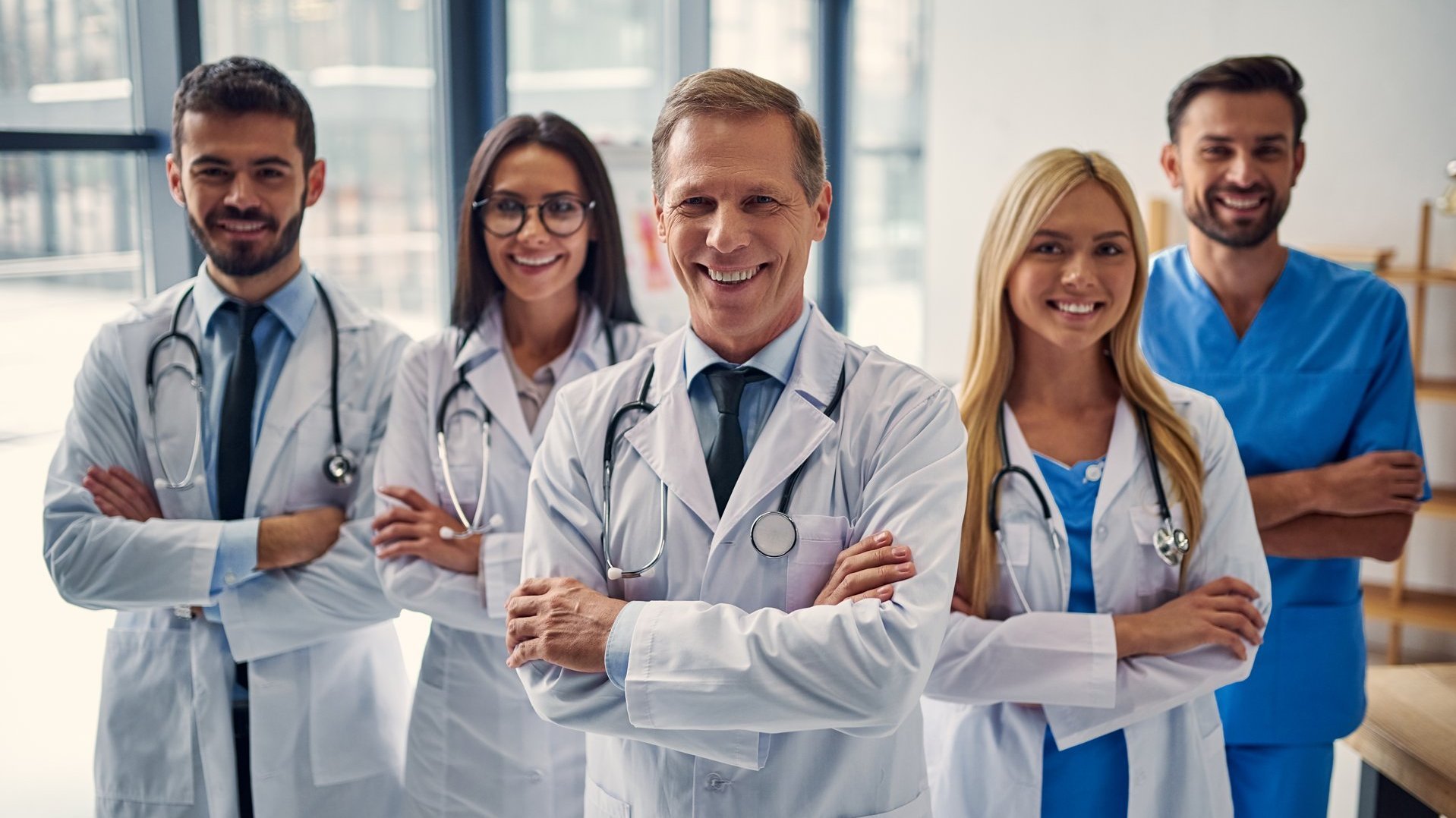 Lächelndes medizinisches Team mit Ärzten und Ärztinnen in weißem Kittel und Pflegekraft in blauer Kleidung im Klinikflur.