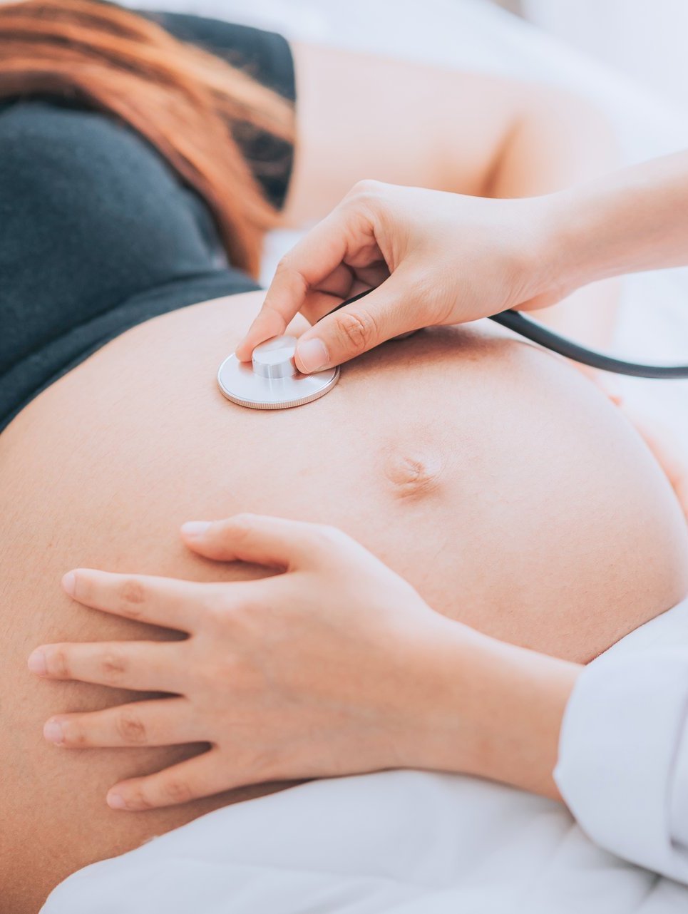 Nahaufnahme eines schwangern Bauches einer Frau. Der Bauch wird von einem Arzt oder einer Ärztin mit einer Hand mit einem Stethoskop untersucht. Mit der anderen Hand wird der Bauch abgetastet.