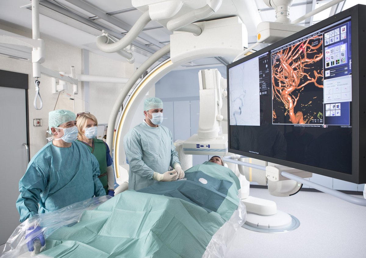 Ärzteteam in einem hochmodernen Katheterlabor führt eine interventionelle Gefäßbehandlung durch, während Angiografie-Bilder auf einem großen Monitor angezeigt werden.