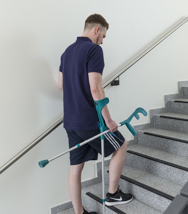 Junger Mann steigt mit Unterarmgehstützen eine Treppe hinauf und hält sich am Geländer fest.