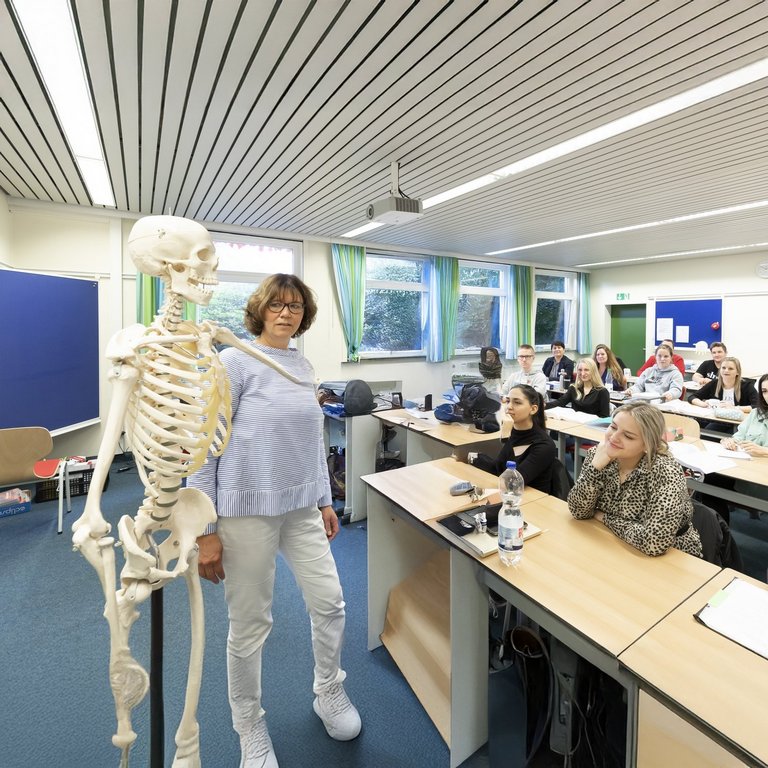 21-09-29-akh-057 Die Szene zeigt einen lebendigen Unterricht im medizinischen Bereich: Eine Lehrkraft erklärt anatomische Zusammenhänge am Skelettmodell, während die Studierenden aufmerksam folgen.