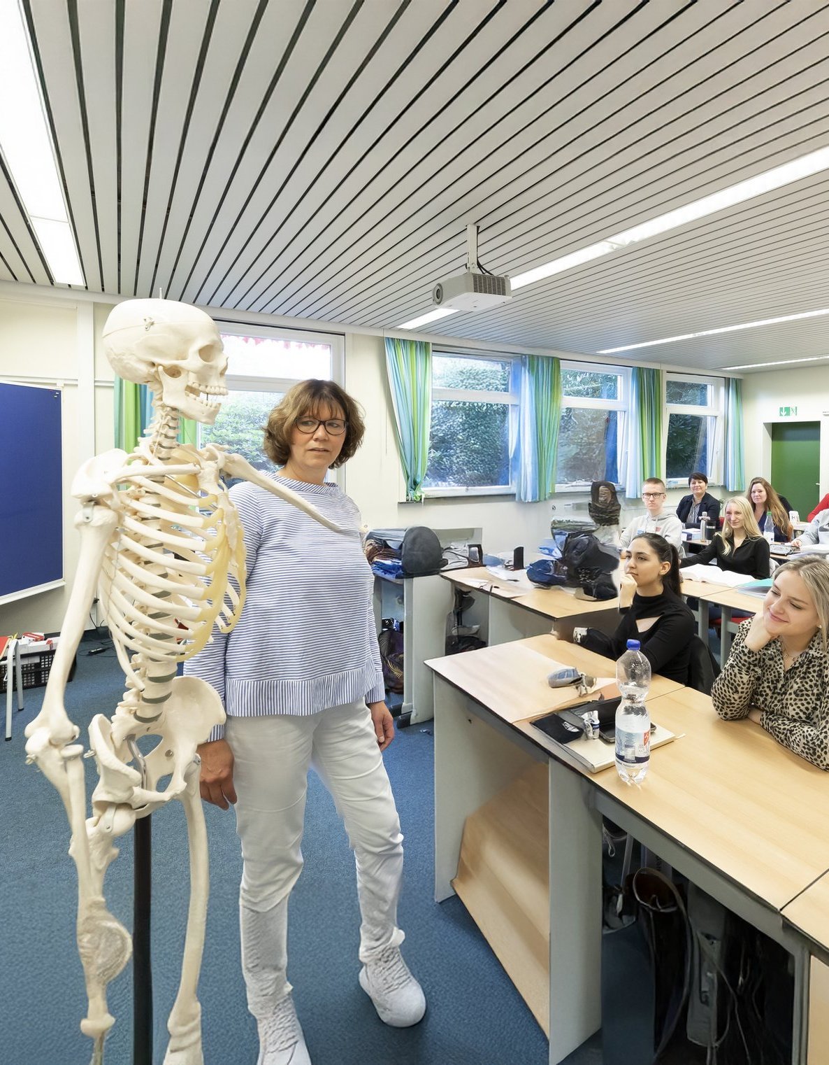 Die Szene zeigt einen lebendigen Unterricht im medizinischen Bereich: Eine Lehrkraft erklärt anatomische Zusammenhänge am Skelettmodell, während die Studierenden aufmerksam folgen.