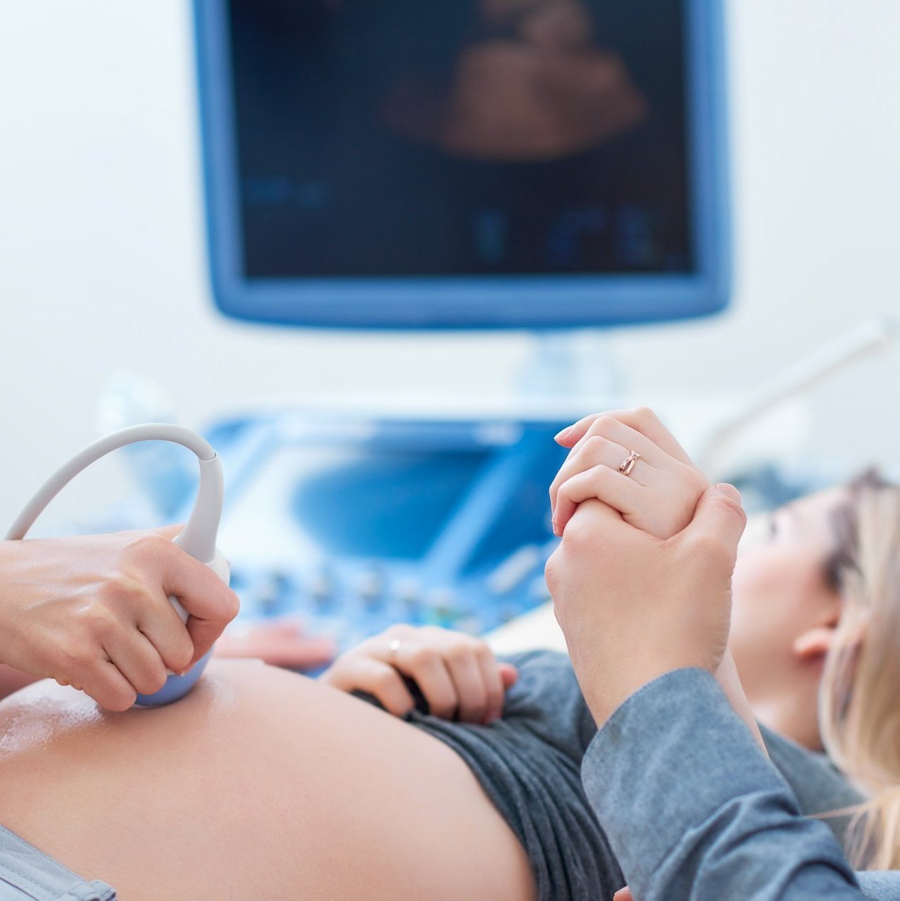 Mit einem Ultraschallgerät wird der Bauch einer schwangern Frau untersucht. Eine weitere Person hält die Hand der Frau. Die Frau blickt in Richtung des Monitors im Hintergrund.